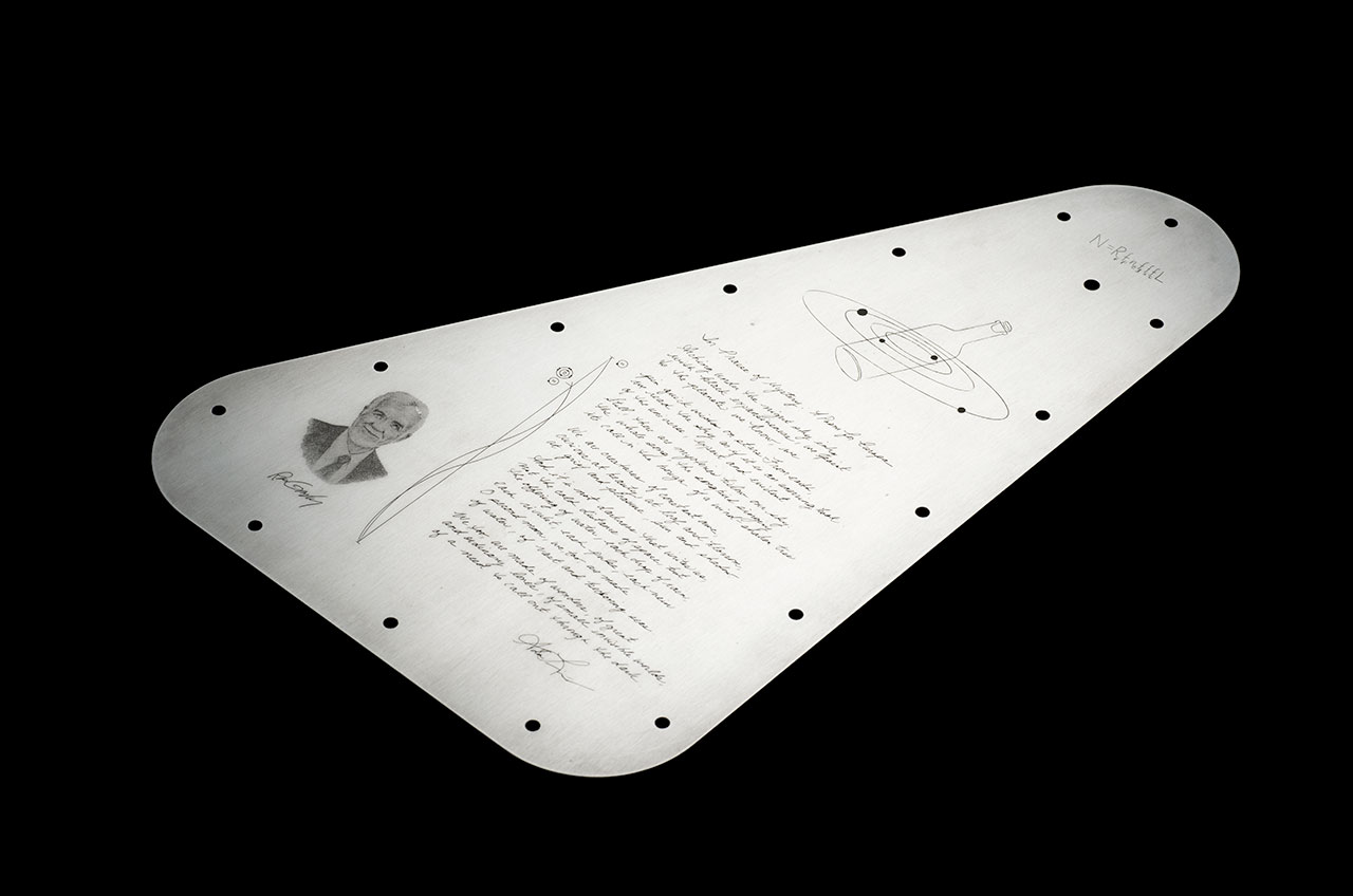 a triangular, silver metal plate with a poem etched on its surface rests against a black background