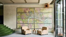 A hotel lobby with a wall-wide, multi-framed vintage map in pink and green tones, two velvety camel armchairs, a glassy door, and velvet green-clad steps.