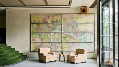 A hotel lobby with a wall-wide, multi-framed vintage map in pink and green tones, two velvety camel armchairs, a glassy door, and velvet green-clad steps.