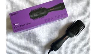 The Amika Blow Dryer Brush with its box against a textured white fabric background