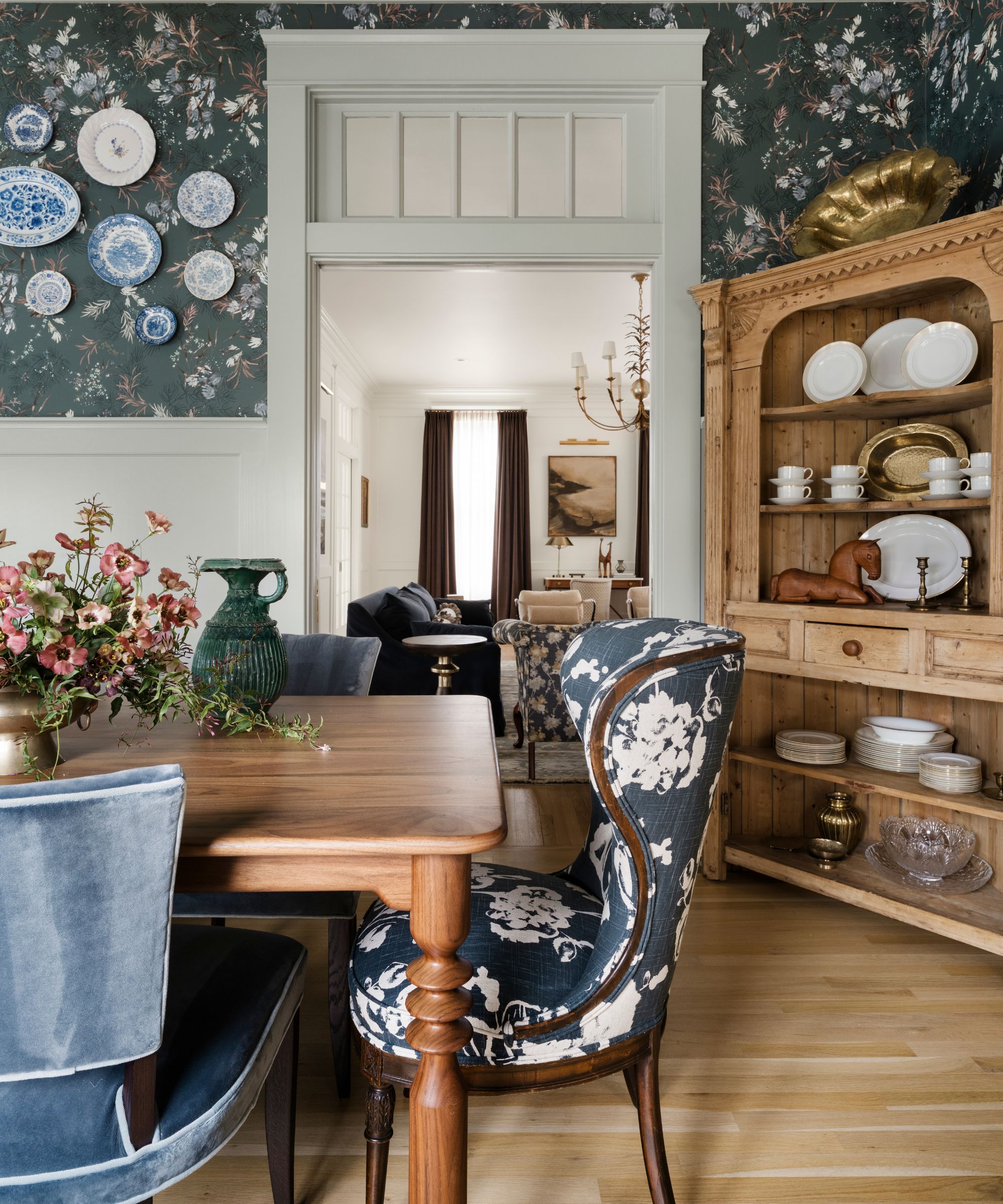 a formal dining room with navy floral wallpaper, original beadboard and millwork, wooden dining table and hutch with a mix of blue floral and velvet chairs