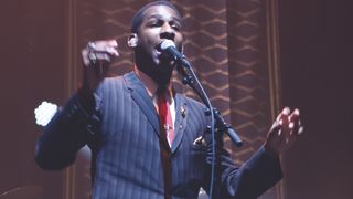 Leon Bridges in a blues, striped suit, white shirt and red tie. singing into a microphone on stage.