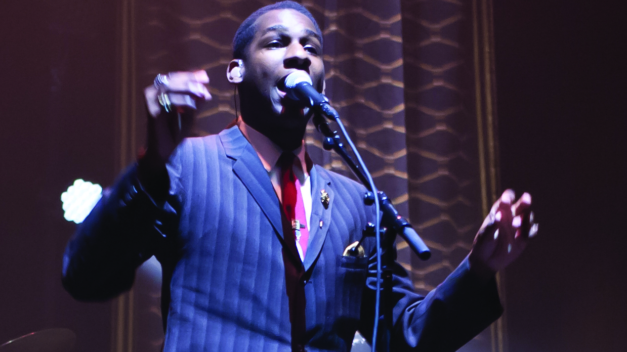 Leon Bridges in a blues, striped suit, white shirt and red tie. singing into a microphone on stage.