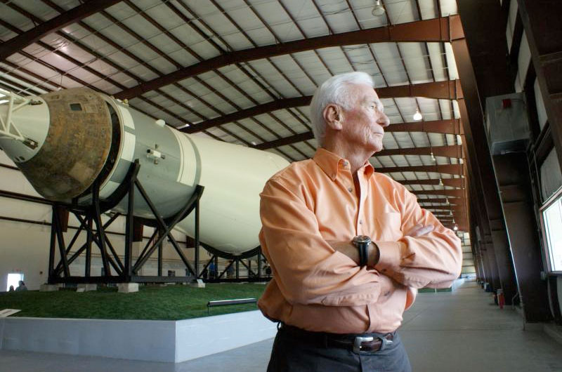 Eugene Cernan, in an orange shirt, crosses his arms in front of part of a spaceship