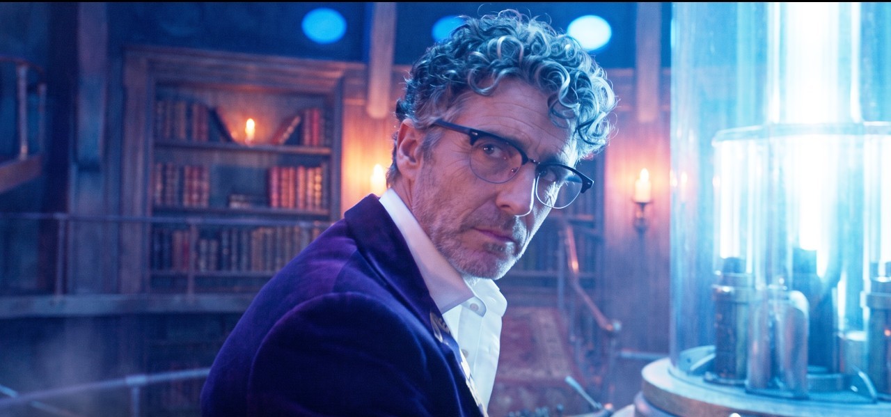 A grey-haired man with curly hair, glasses and a purple velvet jacket looks over his shoulder inside a glowing, book-lined TARDIS interior.