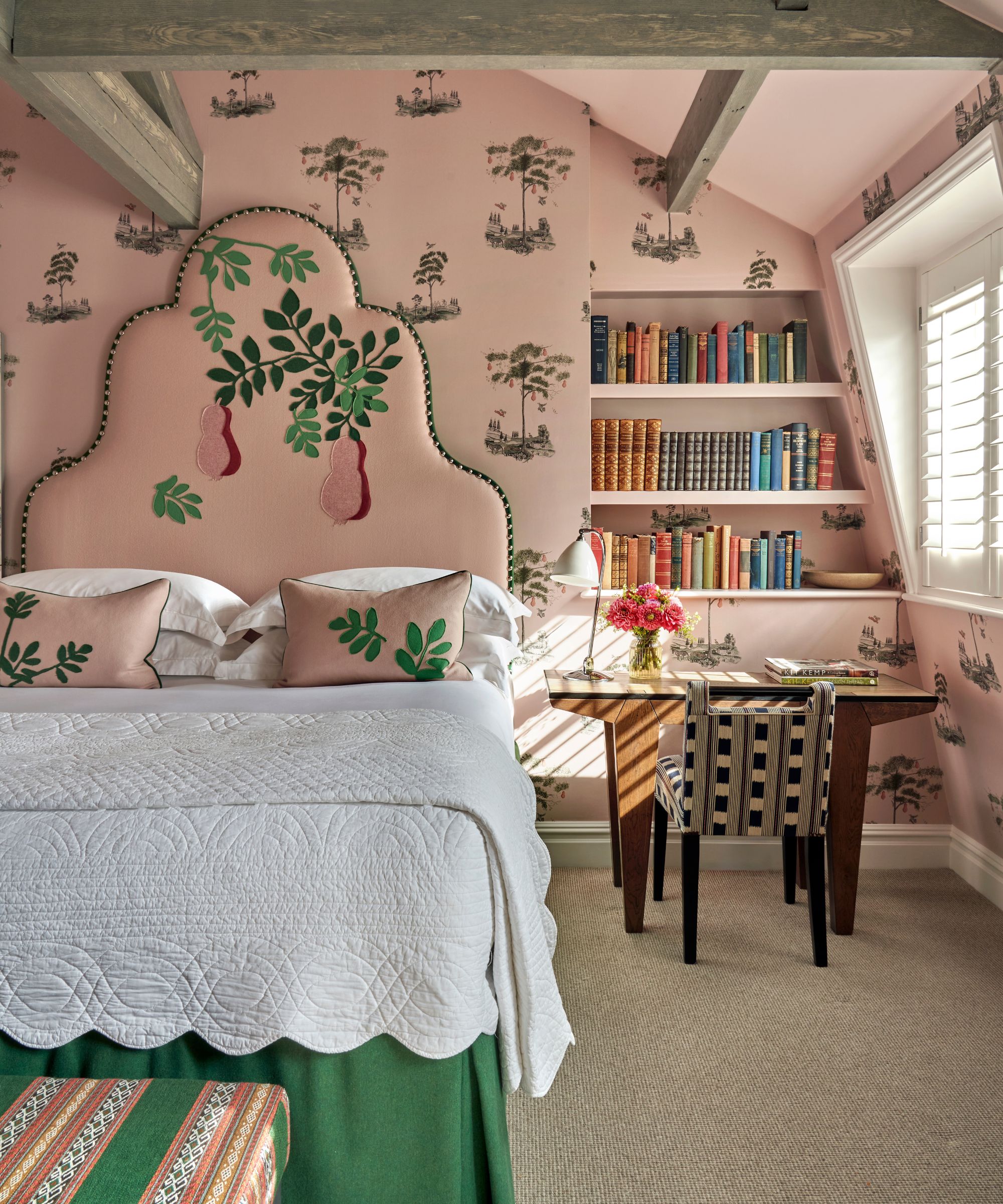pink bedroom with floral wallpaper and a pink embroidered floral headboard with white bed linens, green bed skirt and upholstered ottoman