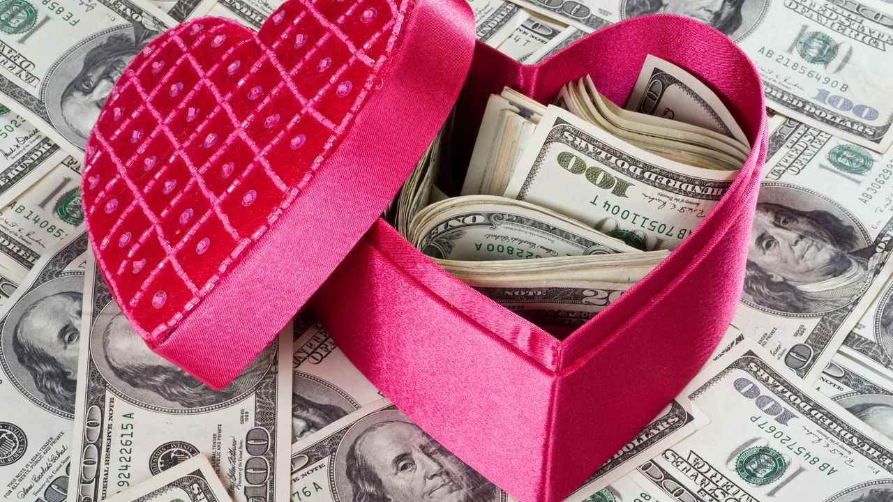 A pink heart-shaped gift box holds several hundred dollars while sitting on top of more cash.