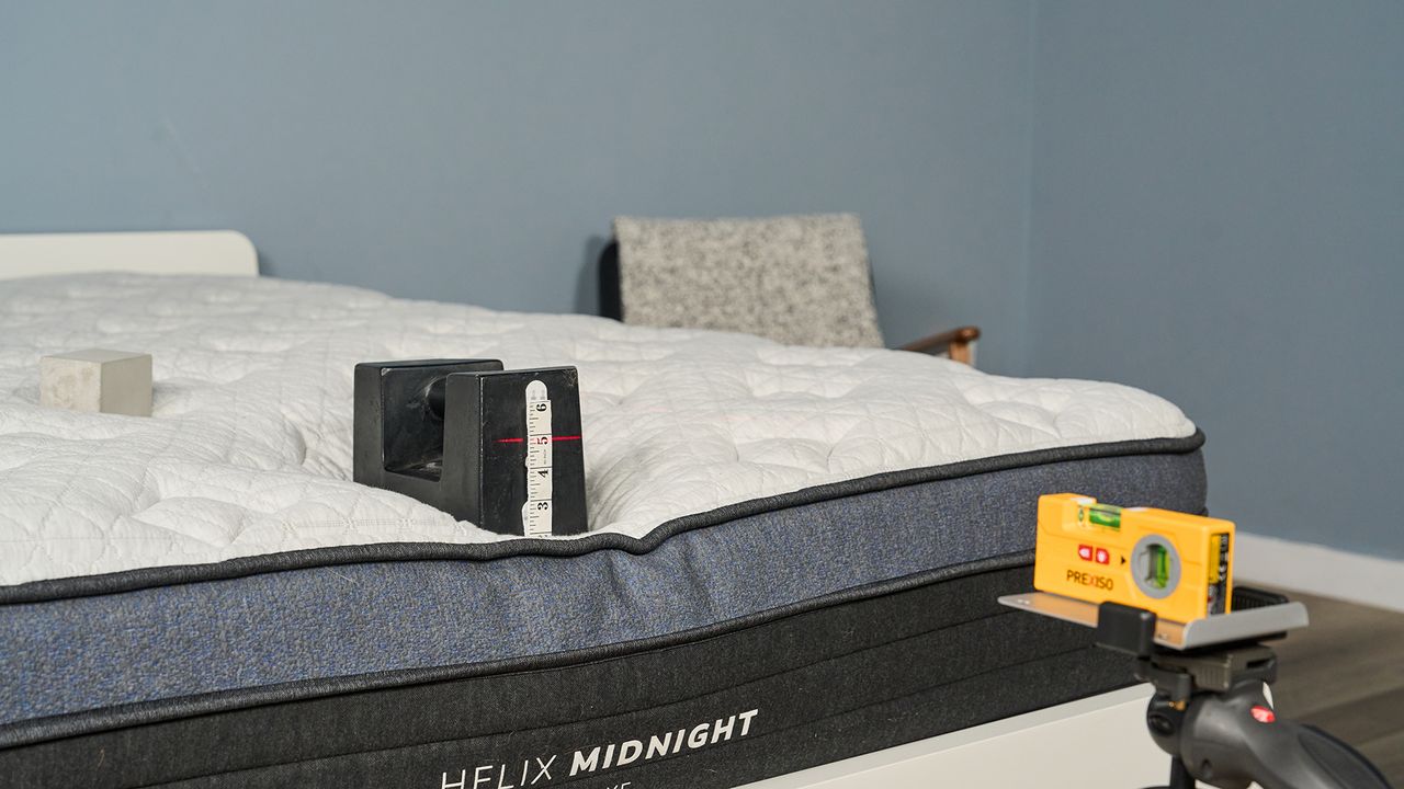 A close up of a 25kg weight balanced on the edge of the Helix Midnight Luxe mattress in our sleep studio. A laser is pointed at the side of the weight where a tape measure is placed.
