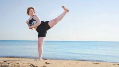 A woman does a kickboxing kick on the beach.