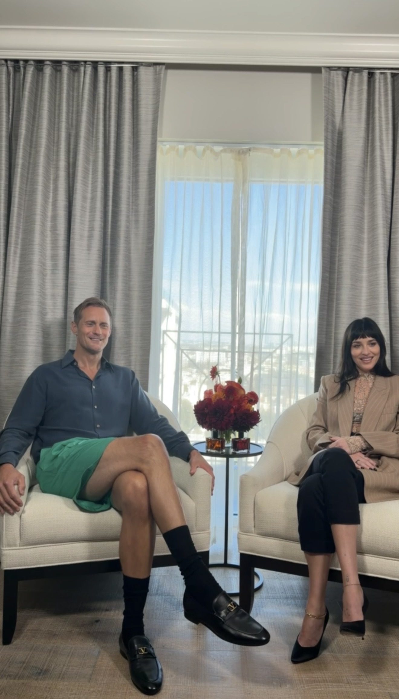 Alexander Skarsg&amp;aring;rd and Dakota Johnson sitting in cream chairs facing the camera