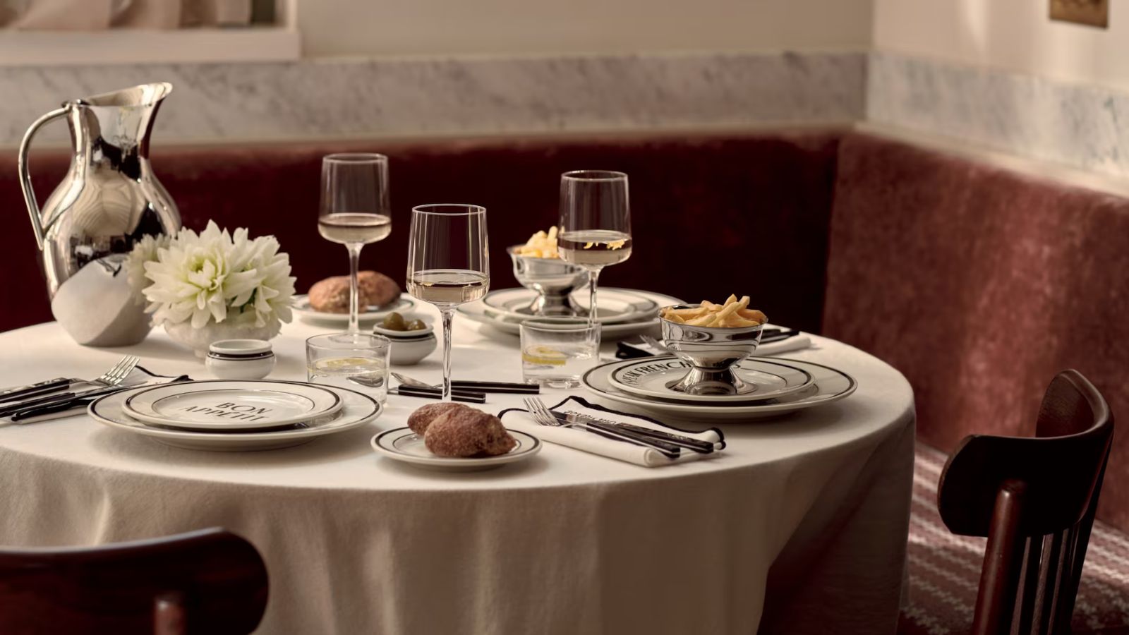 A bistro table with a white tablecloth, black-rimmed plates, three wine glasses and a stainless steel pitcher