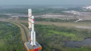 a white rocket rolls out to its launch pad on a track that passes greenery and a small lake