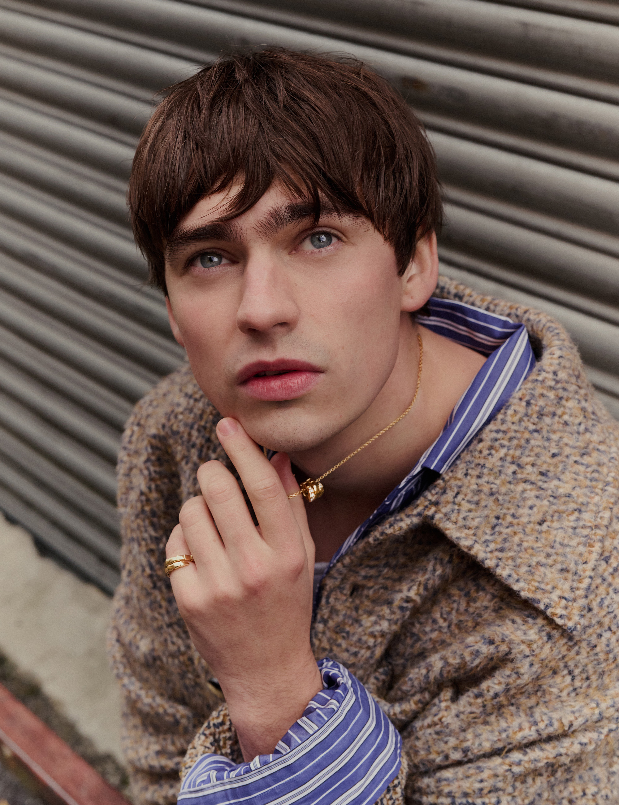 Spike Fearn looks up at the sky while playing with his gold Bulgari necklace. He is wearing a marbled tweed coat over a blue and white stripe button-down shirt.