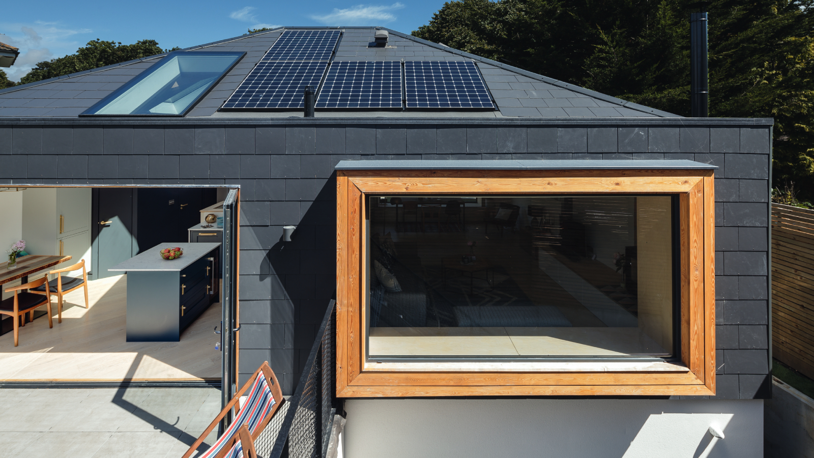 modern new build with solar panels on black roof with large picture window below framed in wood and view through open patio doors into kitchen diner