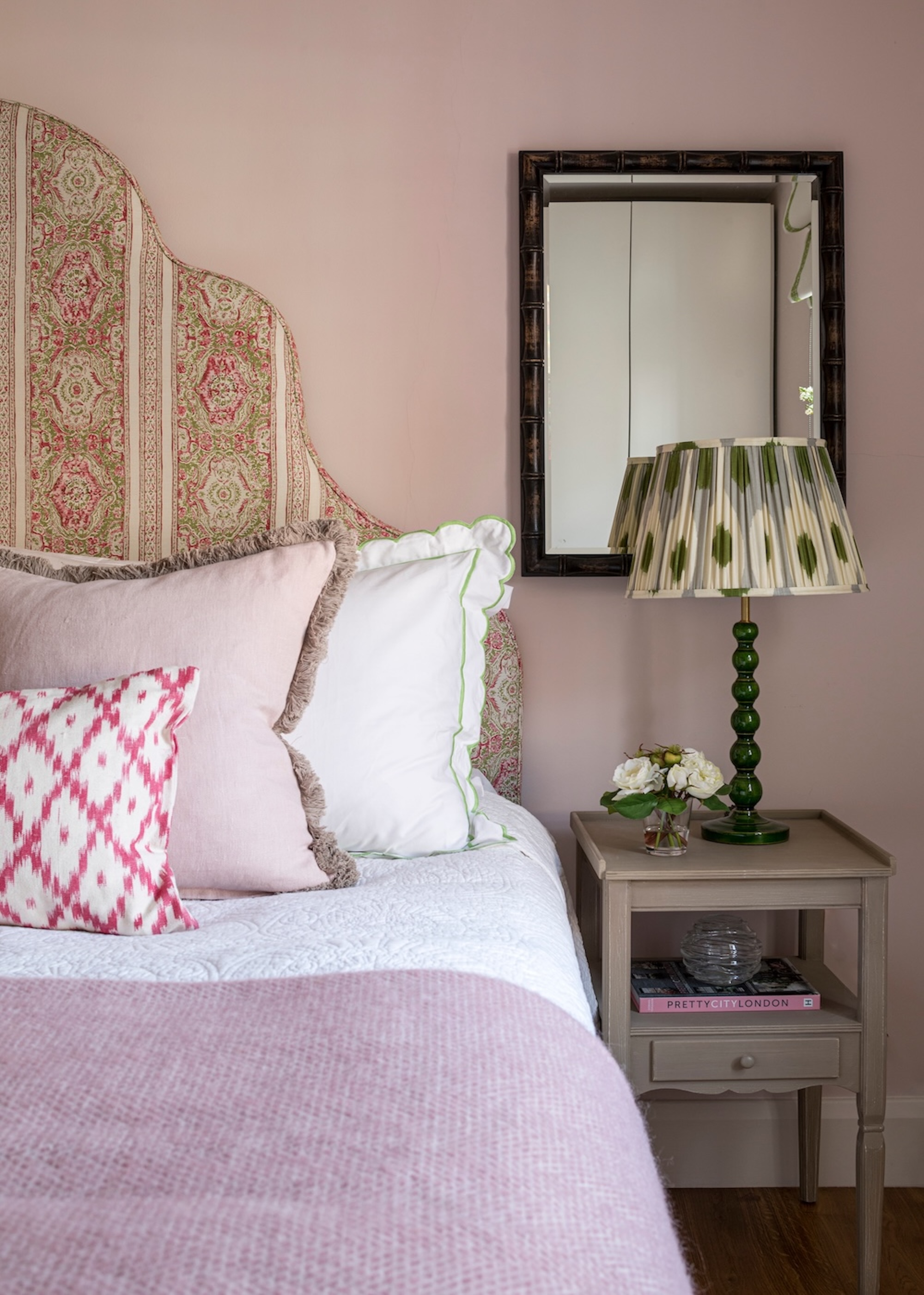 A bedroom with a view of the side of the bed that has a side table next to it and a mirror above it. On the side table is also a lamp.