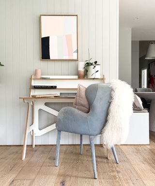 A home office with light gray wall paneling, a framed pastel wall art print, a white wooden desk with three tiers of shelving with plants and books on, and a light gray velvet chair with a white faux fur throw