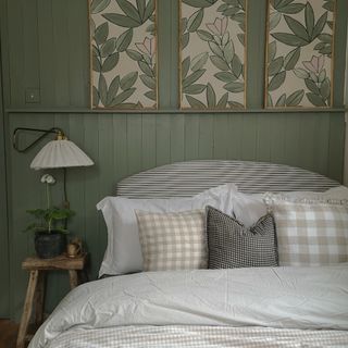 A wood-panelled bedroom painted in sage green with three botanical artworks above the bed with gingham bed linen