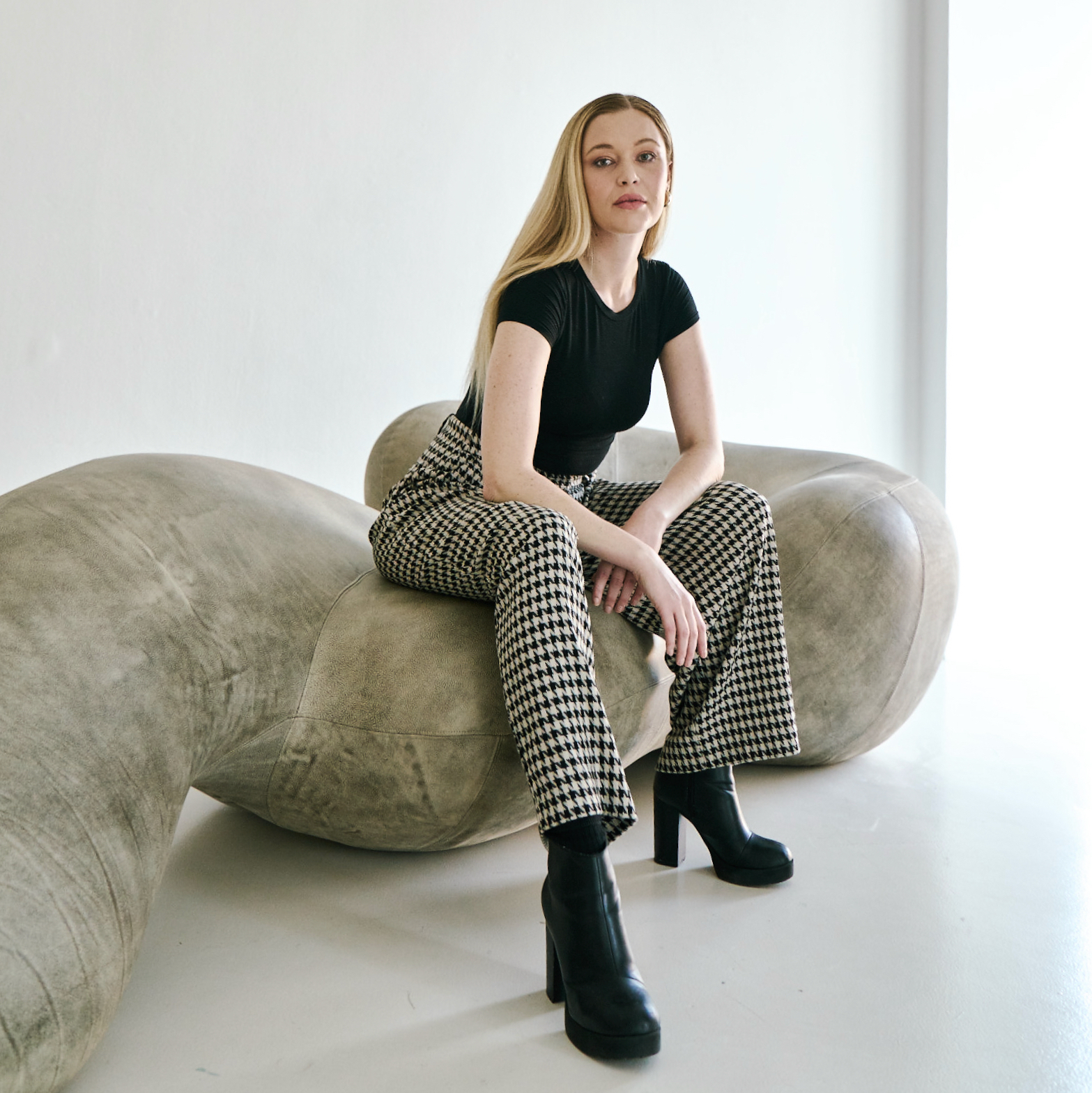 A young woman dressed in a black T-shirt, checkered white and black trousers, and black high-heel boots sits on a large, winding, stone-like sculpture in a white, naturally lit gallery.