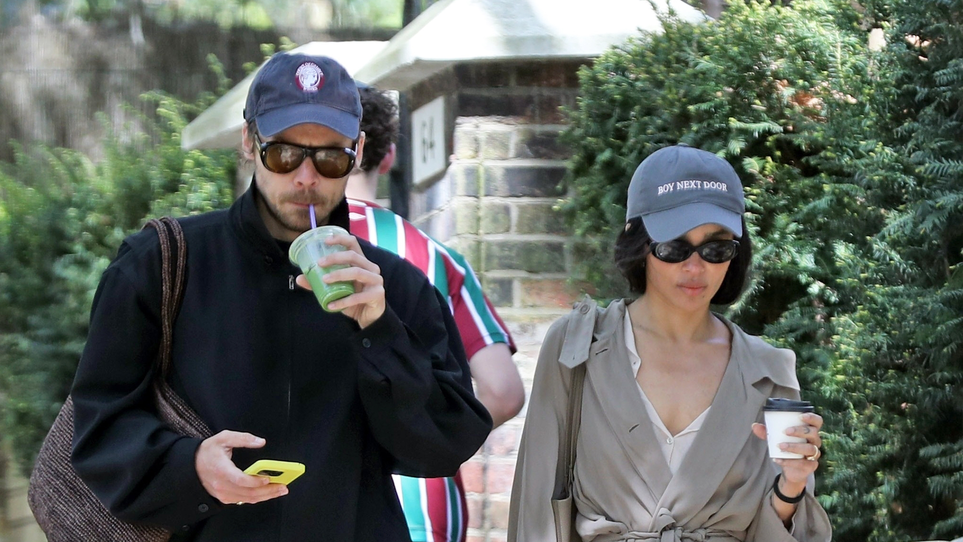 Zoe Kravitz wearing a navy baseball cap, tan trench coat, white blouse, black pants, and The Row soft loafers