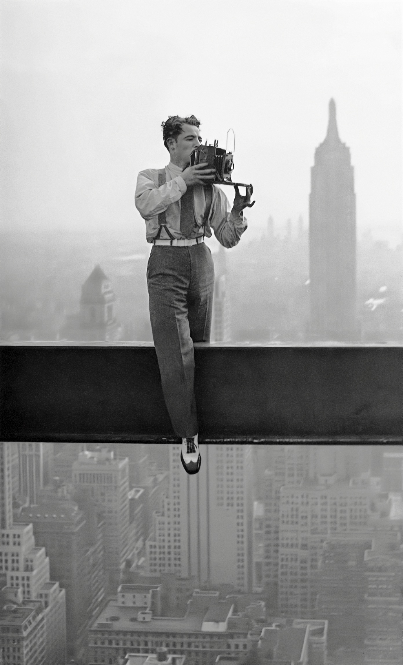 A young photographer in suspenders and two-tone shoes sits astride a narrow steel beam dozens of storeys above Manhattan, raising a large plate camera to his face with the Empire State Building looming in the fog behind him.