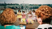 From a behind-the-shoulder perspective of two judges, several young girls in frilly dresses hold numbered signs during a beauty pageant at an outdoor pool.