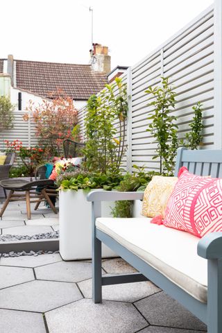 a white garden with hexagonal paving, a white fence with plants growing up it, and black and colorful garden furniture