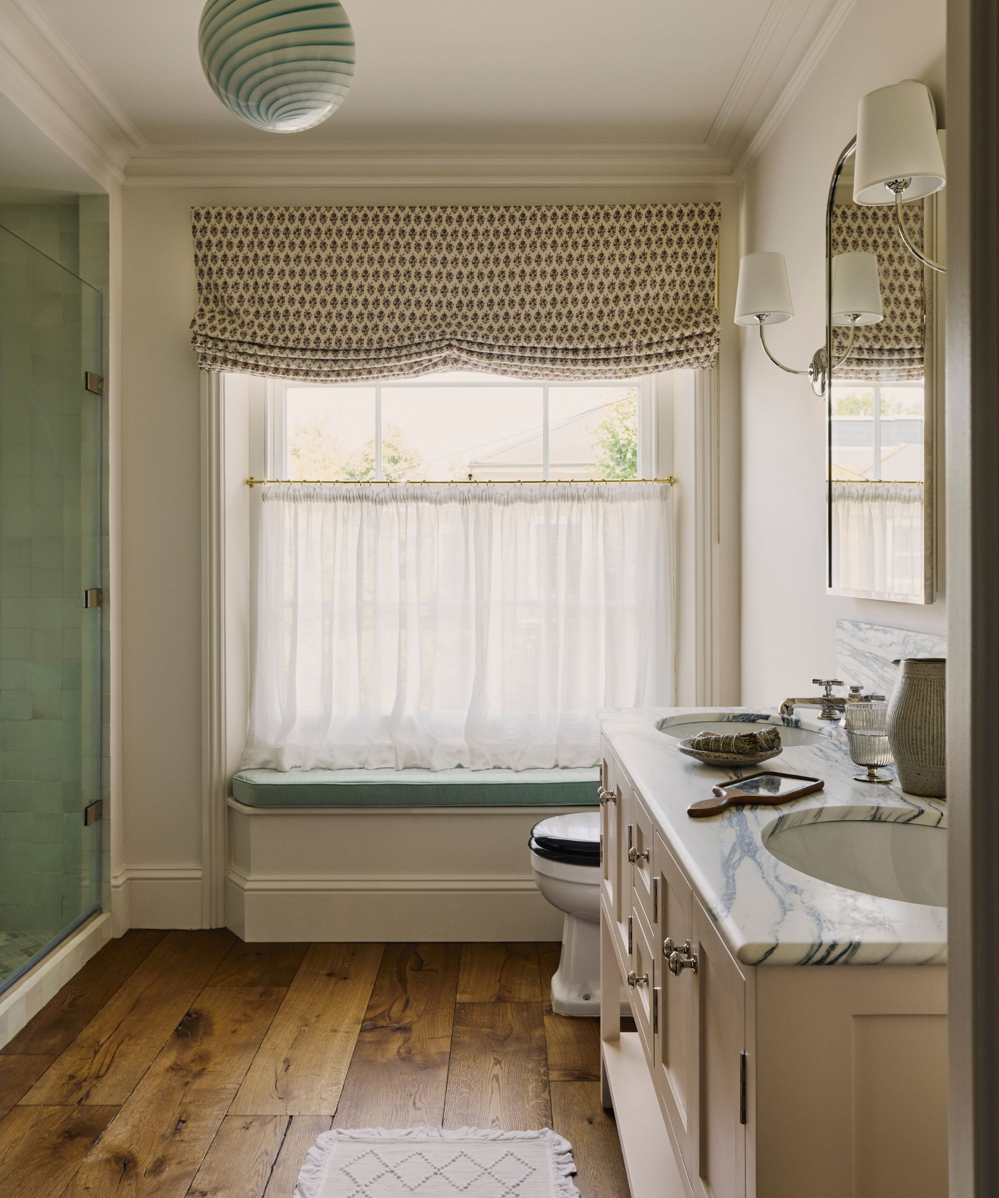 Bathroom with marble vanity unit, wooden floor and large window with blue window seat, floral blind, and sheer cafe curtain