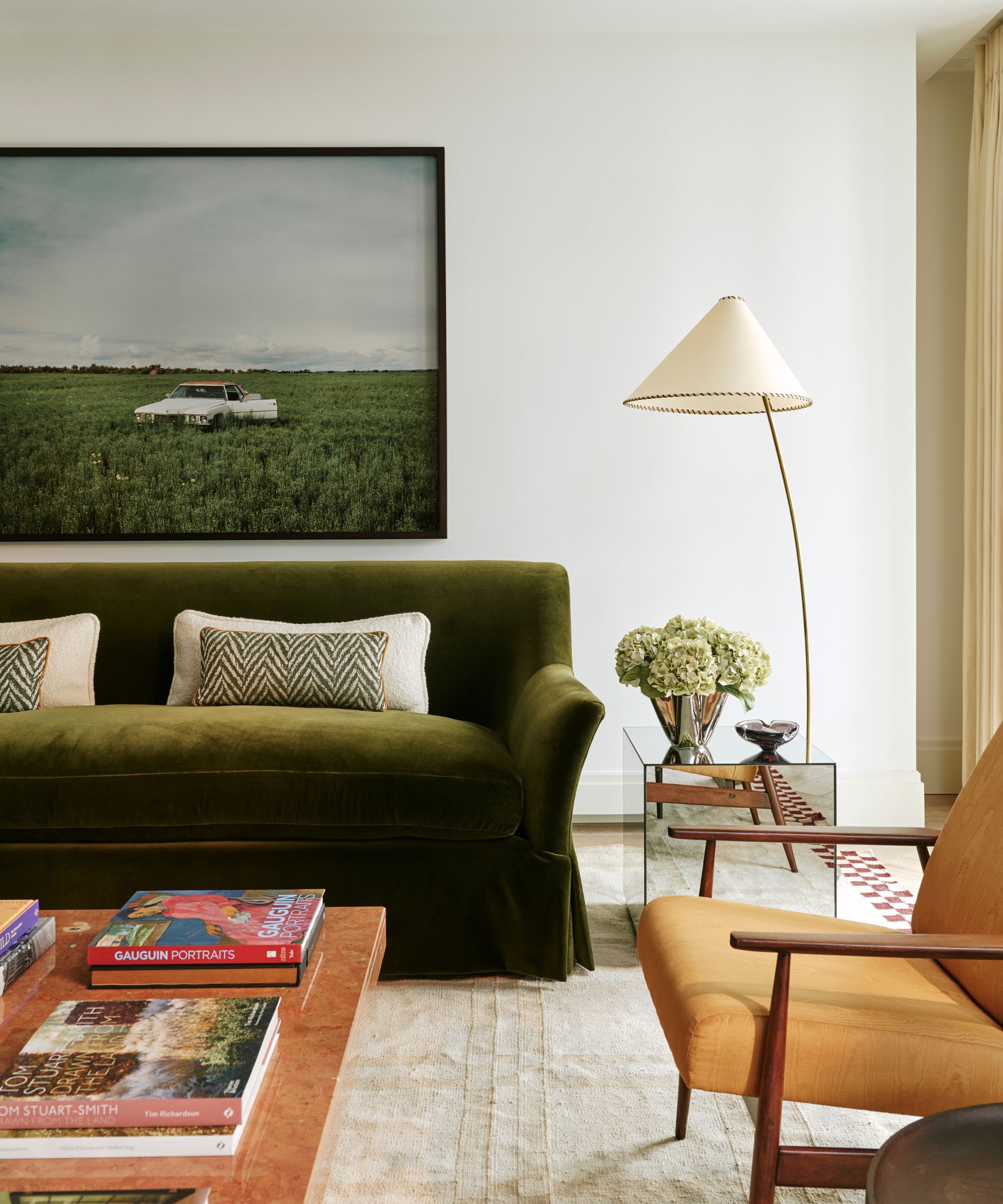 neutral living room with a plush rug, dark green velvet couch, red marble coffee table styled with stacks of books, a wooden and ochre armchair and a floor lamp
