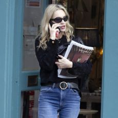 Kate Moss steps out, taking a phone call. She wears blue bootcut jeans with a black jumper, black loafers and a black belt. She carries a stack of magazines in the crook of her arm.