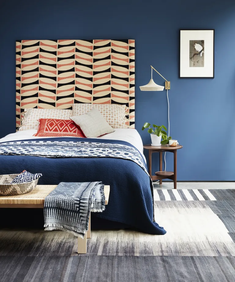 A bedroom with dark blue walls, a patterned orange and blue headboard and a blue and white woven rug