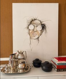 Portrait painting of Joy Moyler on top of a white table, silver serverware, ornaments and books placed in front, wooden walls behind