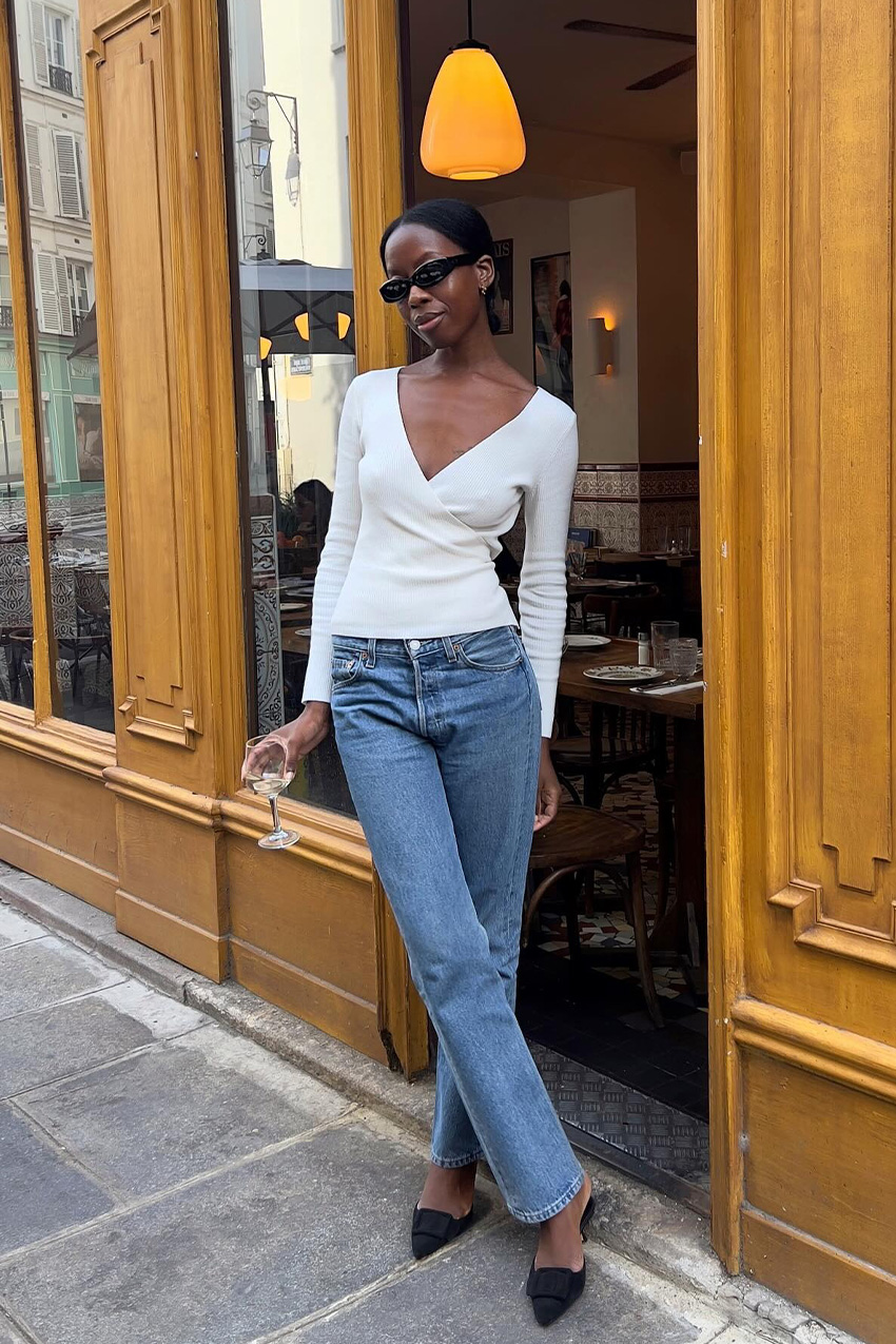 How to wear skinny jeans is shown in a photo of a woman standing outside, leaning against a restaurant doorway, wearing black sunglasses, a white wrap knit top, and low-rise skinny jeans and black pointed mules
