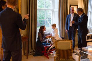 Prince George on a rocking horse in a white robe with Princess Kate kneeling in front of him and Prince William talking to Barack Obama behind