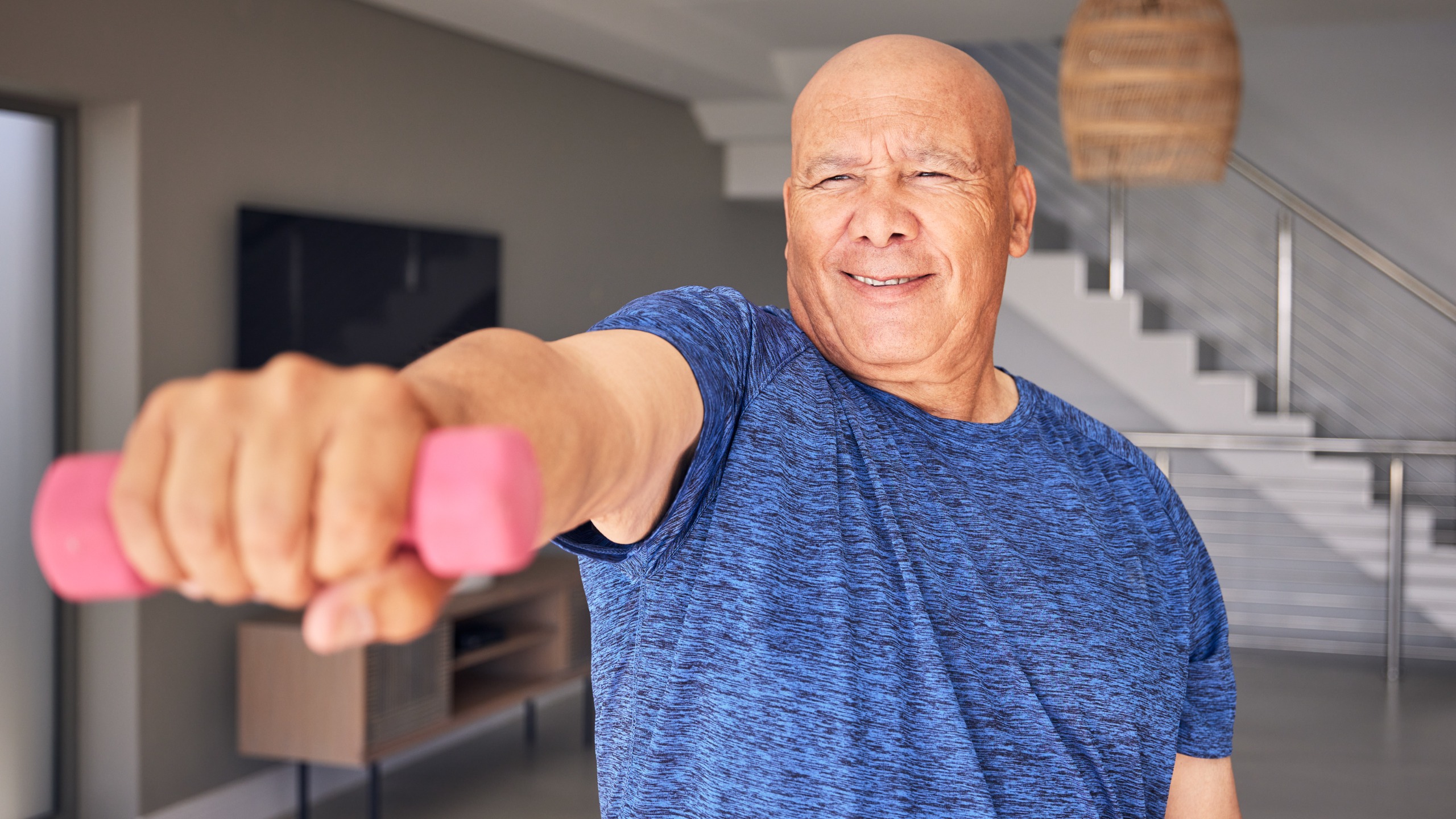 A smiling man in a t-shirt holds a light dumbbell up towards the camera, his arm extended in a straight line from his shoulder.