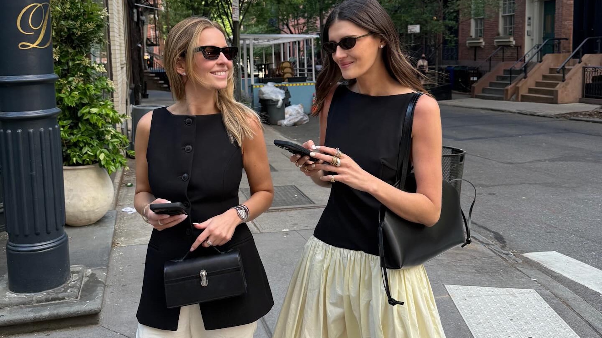 NYC style influencer Coco Schiffer is all smiles with a friend walking at a crosswalk both wearing stylish spring outfit ideas with black sunglasses, a two-tone dress, black vest, black bags, and more