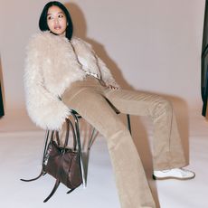 Model sat on a chair wearing a cream fluffy jacket and camel corduroy flared trousers holding a brown handbag sold at Monki