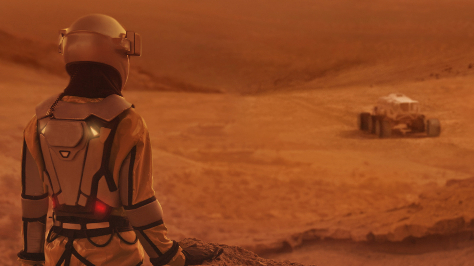 A woman stands to the left of a rover on a red surface