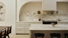 A spacious long off white kitchen with arches and open shelving above shaker cabinetry in a warm neutral