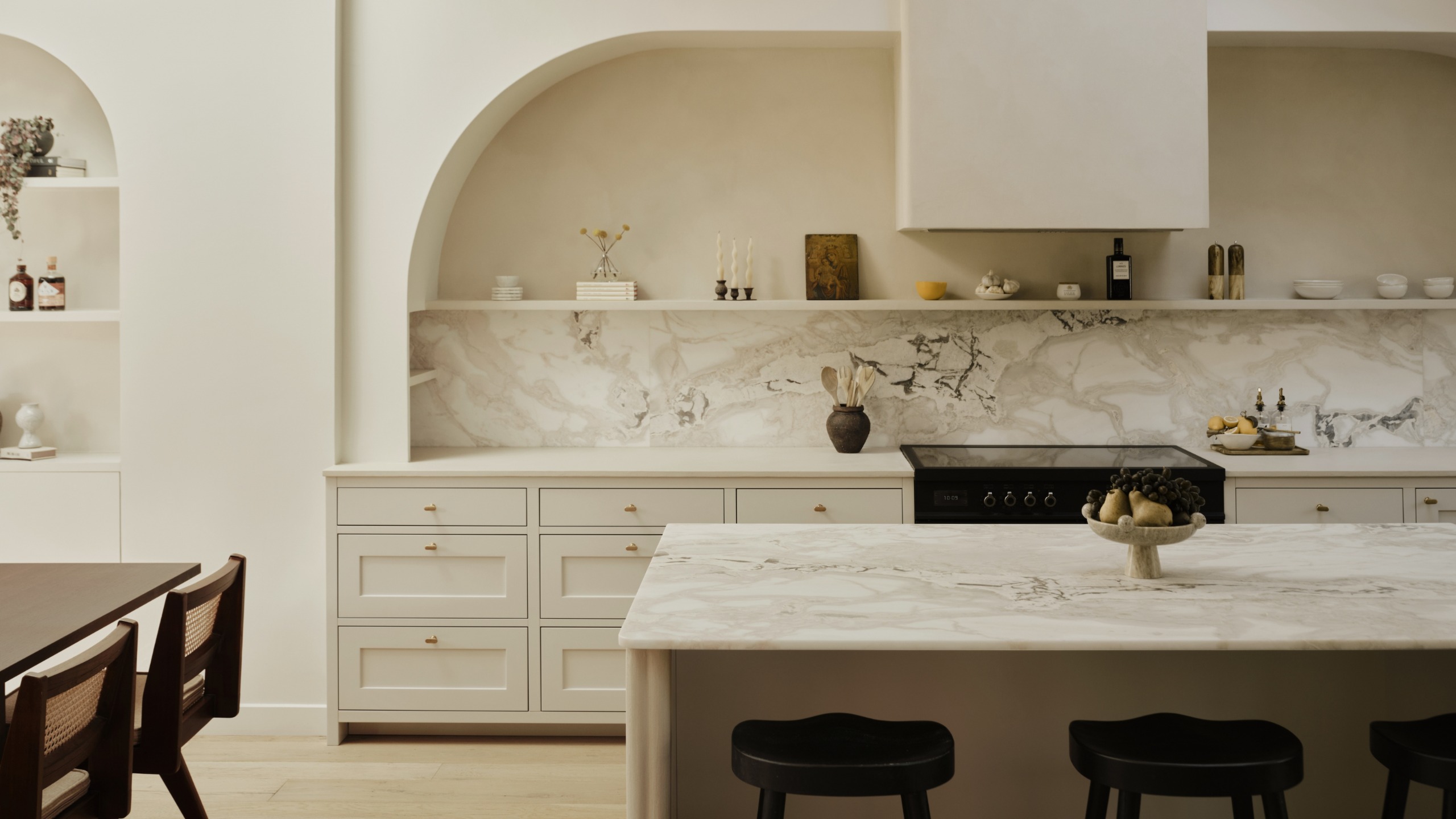 A spacious long off white kitchen with arches and open shelving above shaker cabinetry in a warm neutral