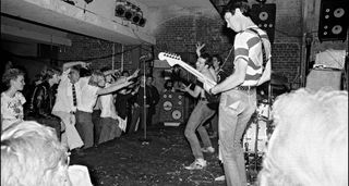 A live shot from the side of the stage as Dead Kennedys play the Mabuhay in 1978. East Bay Ray wears a hooped T-shirt and plays a Strat while a shirtless Jello Biafra works the crowd in the background.