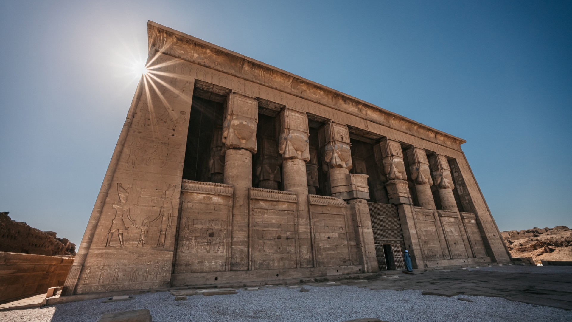 A luz do sol brilha sobre um antigo templo egípcio de arenito, seus relevos esculpidos e colunas imponentes erguendo-se contra um céu azul claro no deserto.