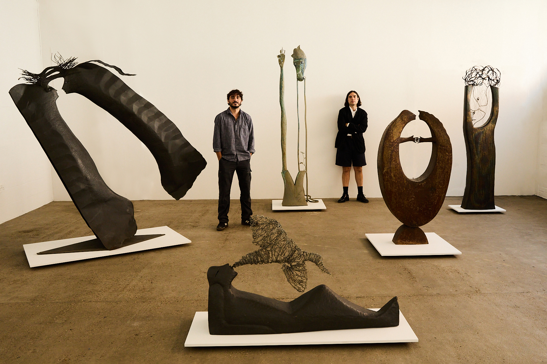 Two young men dressed in casual chic, dark clothing stand proudly in an industrial gallery space surrounded by iron-made, large-scale sculptures modeled after whimsical human silhouettes.