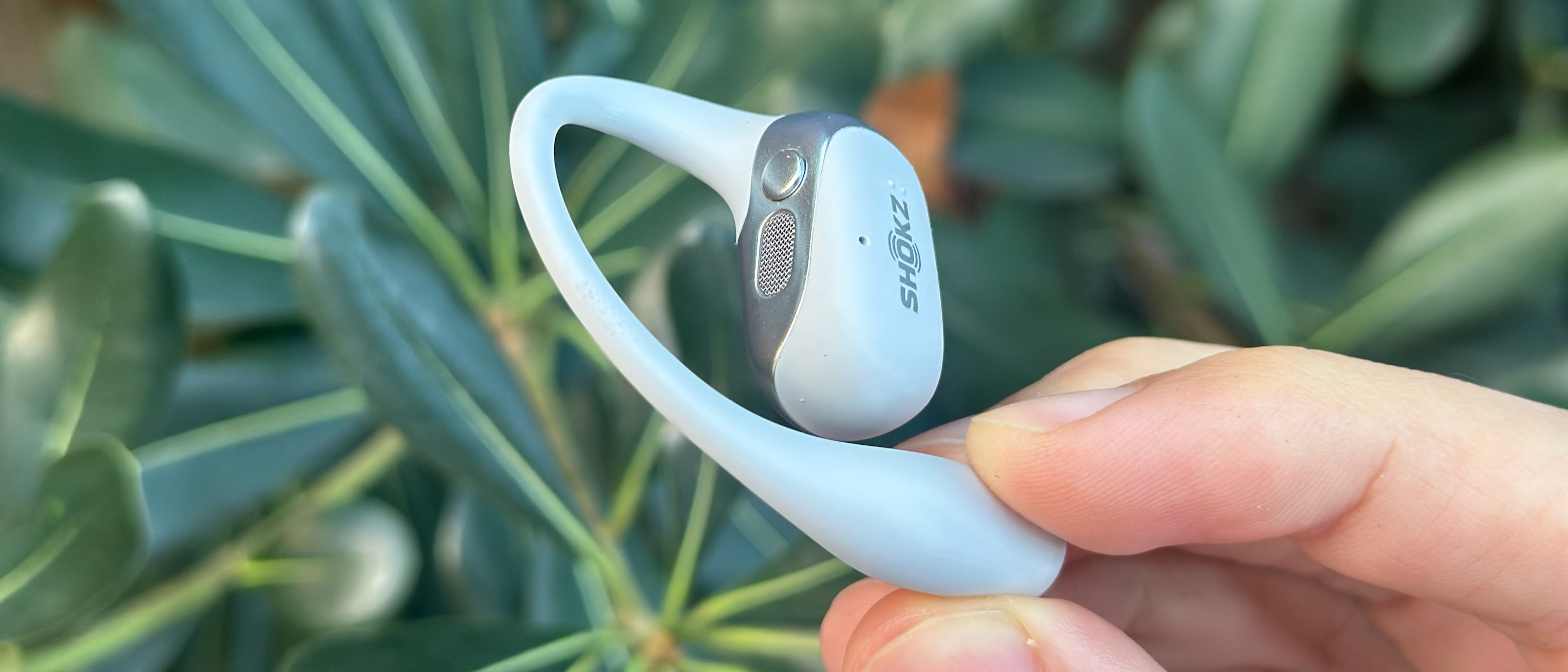A Shokz OpenFit 2 Plus earbud held by the ear hook tip above some green foliage, angled to emphasize the top speaker grill and physical button.