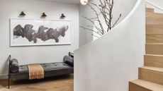 white entryway with curved timber staircase, leather day bed bench seat, an artwork lit by three wall sconces