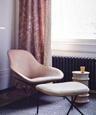 Armchair and footstool in a corner with wooden flooring, grey rug and black radiator in a corner near a large sash window.
