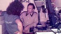 Dave and Philip Sharkey talk in the cramped darkroom at Passport Photo Service in 1978, with a Kodak Versamat processing machine and a Beseler enlarger visible among the equipment around them.