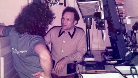Dave and Philip Sharkey talk in the cramped darkroom at Passport Photo Service in 1978, with a Kodak Versamat processing machine and a Beseler enlarger visible among the equipment around them.