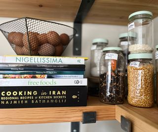 Assorted jars on a thick wooden shelf with white labels and green painted lids next to a stack of recipe books and mesh basket of eggs