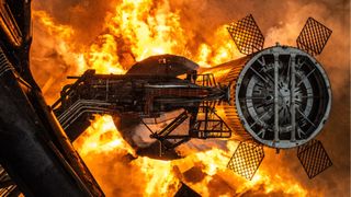fire erupts from below a giant chrome cylinder with four grid fins, next to a tower.