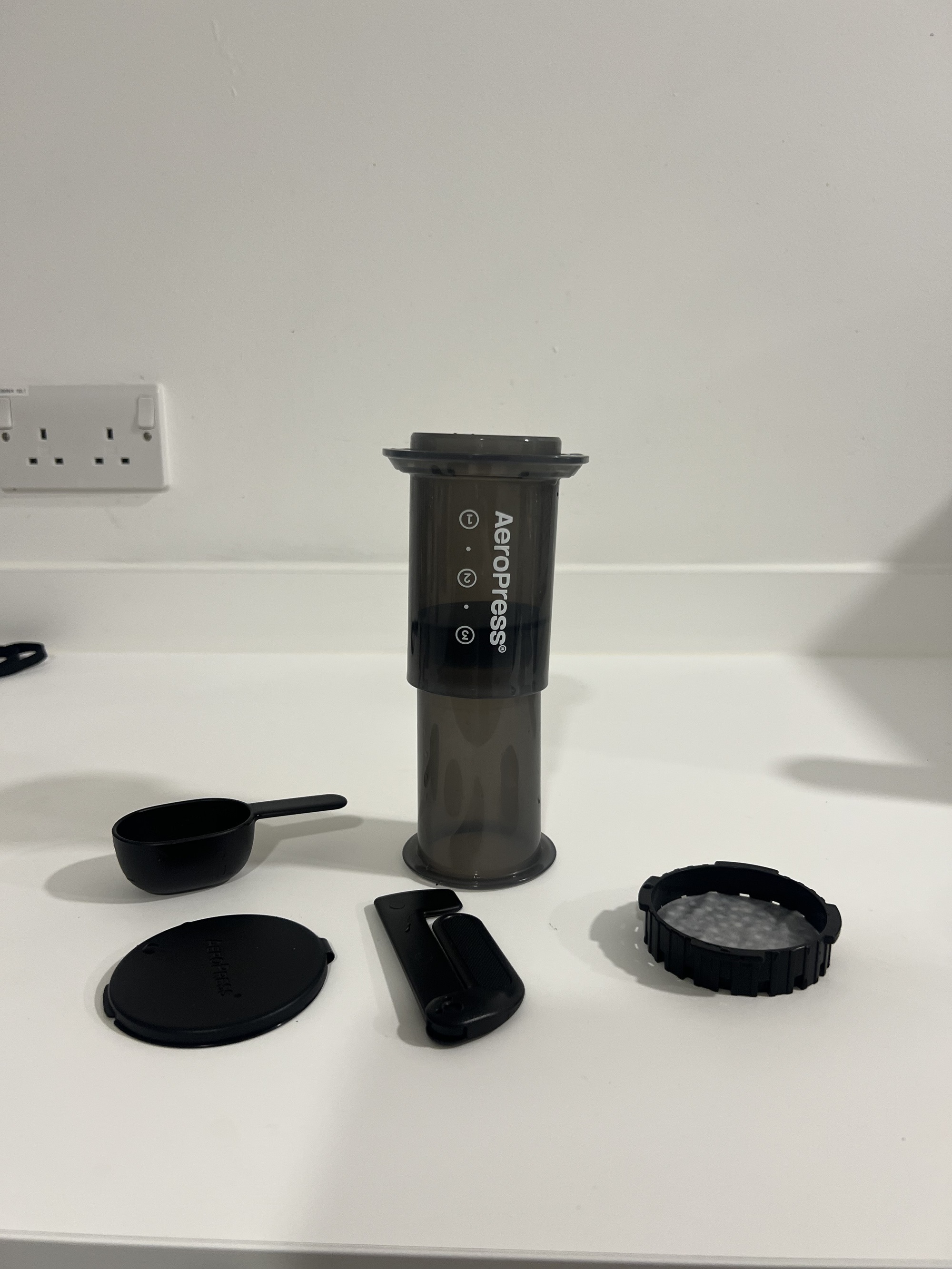 Image of a black AeroPress coffee maker and four of the corresponding coffee making accessories on a white kitchen countertop.